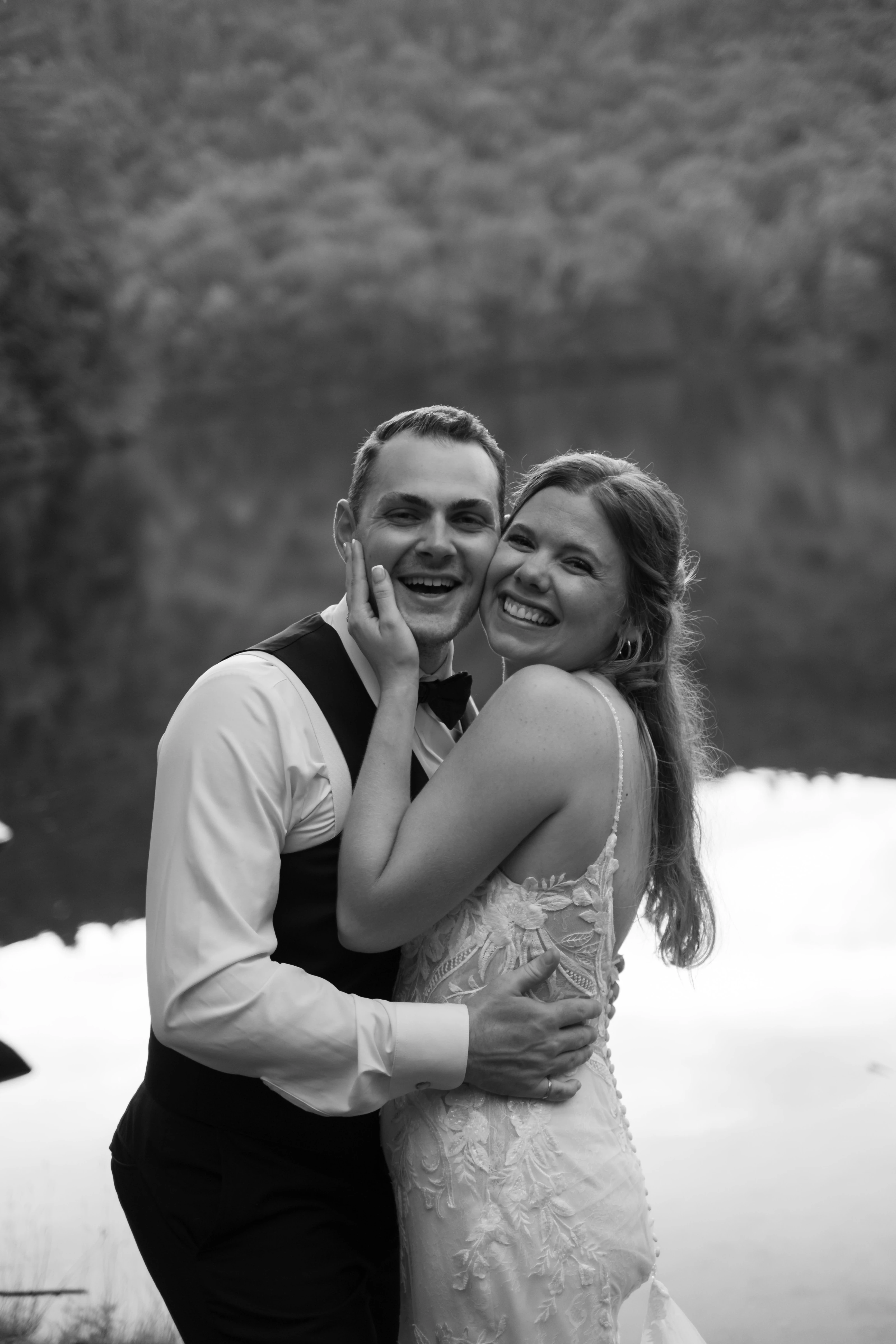 The newlyweds laugh cheek to cheek beside the water in a black-and-white photograph