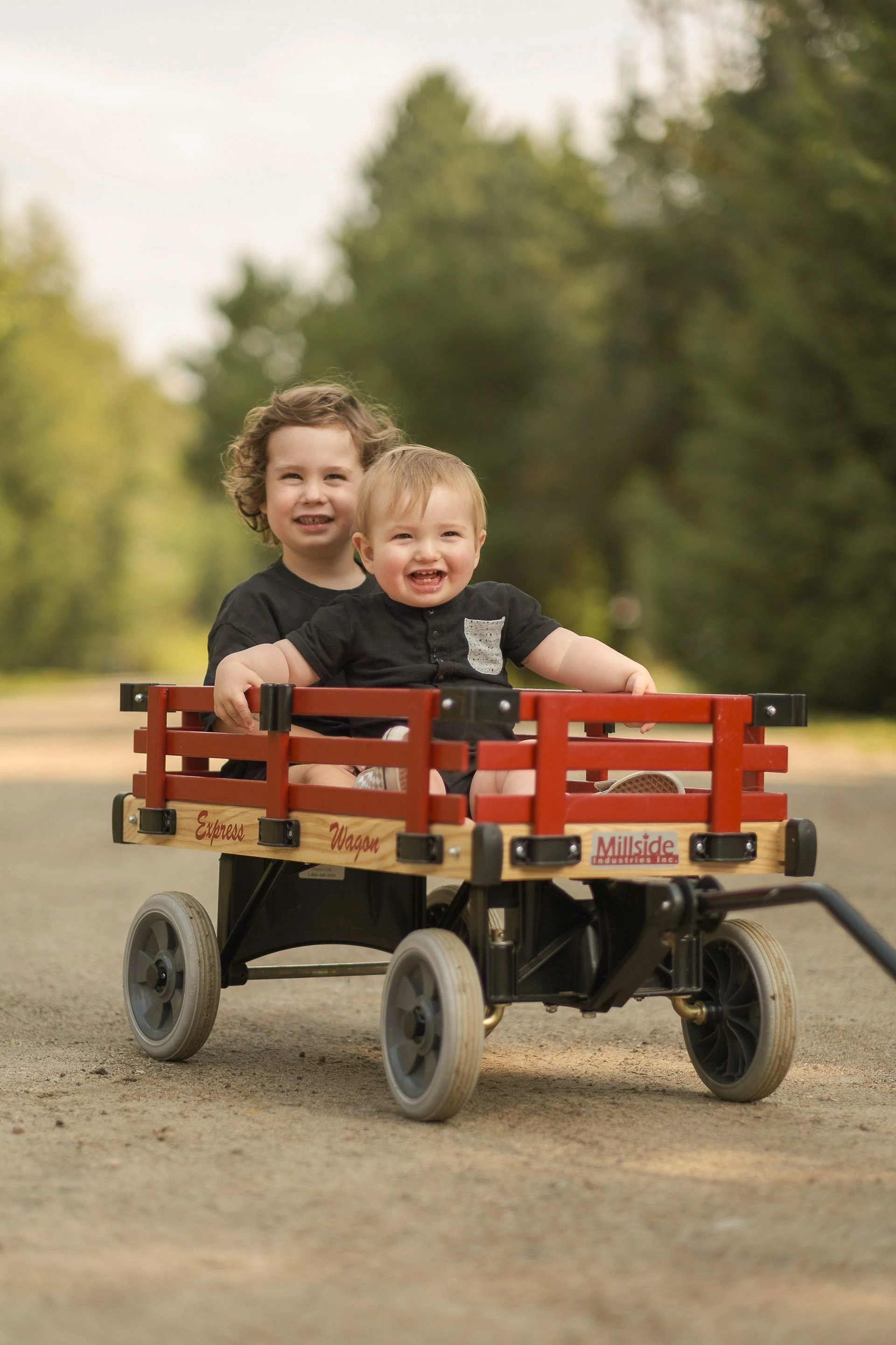 Two young boys laugh in a red wagon on a tree-lined path