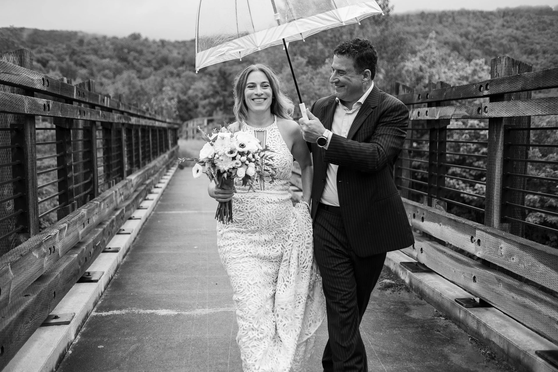 The newlyweds walk hand in hand across a lawn; the bride wears a lace dress and veil and carries a bouquet in front of willows and tall golden grass