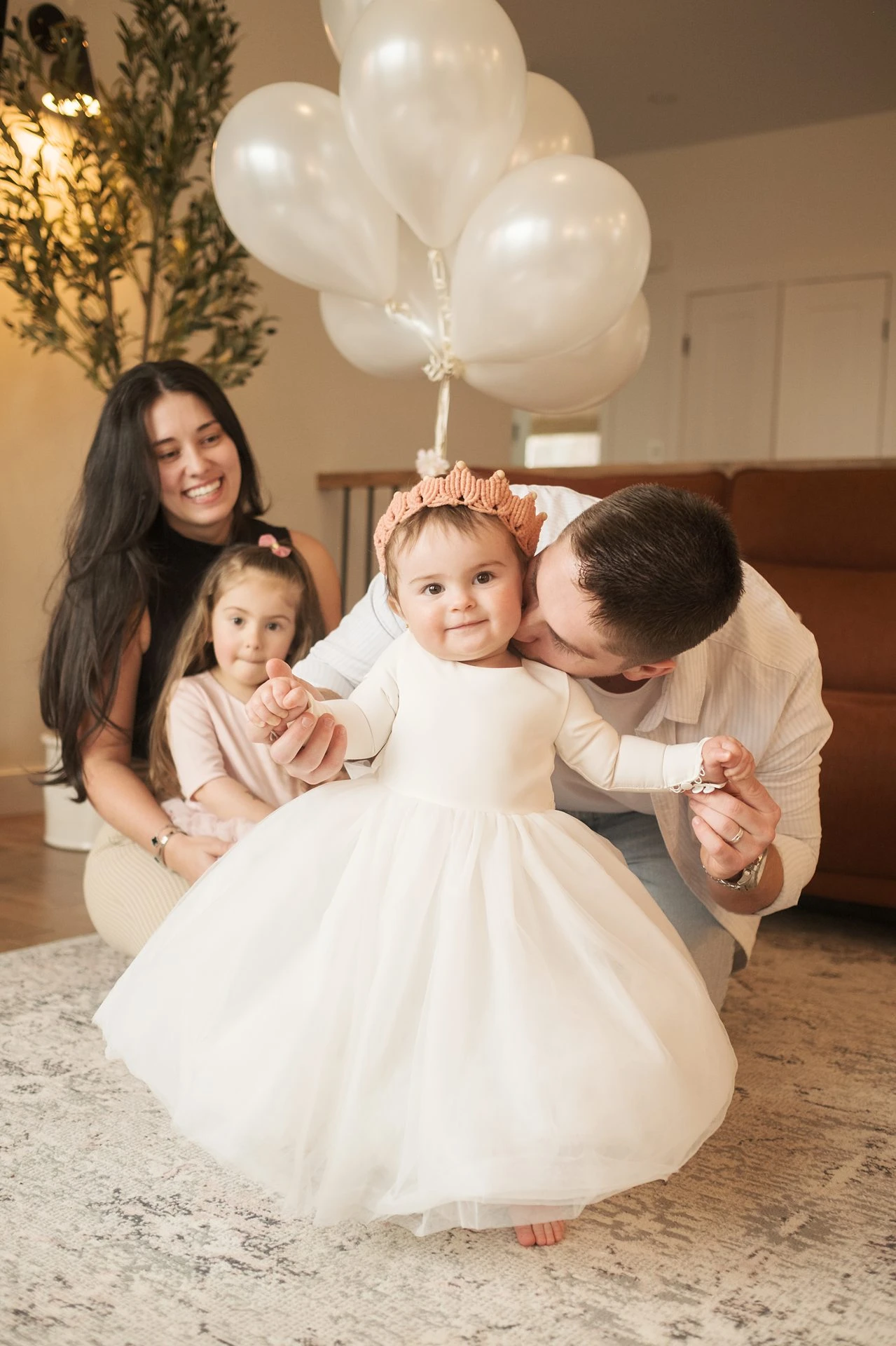 A baby in a white dress and fabric crown with her parents and older sister
